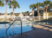 Thumbnail 10 of 15 - Community pool with sundeck, lounge chairs, with lake view surrounded by white metal fence with palm trees and building exteriors in the background