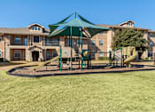 Thumbnail 21 of 27 - A playground with a green canopy and a yellow slide is in front of apartment buildings.