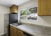 Thumbnail 11 of 18 - Spacious Kitchen with Wooden Cabinetry, Refrigerator, and Wide Windows Looking in Front Space at Park 210 Apartment Homes, Edmonds, WA, 98026