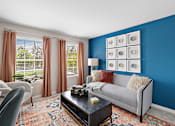 Thumbnail 11 of 24 - A living room with a grey couch, a coffee table, and a window with curtains at Briarcliff Village, Michigan, 48390