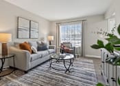 Thumbnail 4 of 16 - A living room with a grey couch, a glass coffee table, and a large window with blinds.