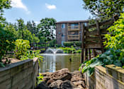 Thumbnail 1 of 31 - A wooden fence borders a pond with a waterfall and a building in the background.