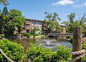 Thumbnail 4 of 31 - A fountain in the middle of a pond surrounded by greenery and a building in the background.