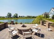 Thumbnail 36 of 73 - A patio with a fire pit and chairs overlooks a lake.