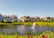 Thumbnail 39 of 73 - A serene lakeside scene with a fountain and houses in the background.