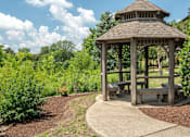 Thumbnail 11 of 23 - A wooden gazebo with a brown roof is surrounded by greenery and has a circular concrete walkway around it.