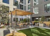 Thumbnail 14 of 30 - A patio with a table and chairs is surrounded by a grassy area and a building with large windows.