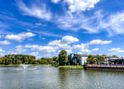 Thumbnail 51 of 51 - A serene lake with a fountain in the middle, surrounded by trees and buildings under a clear blue sky.
