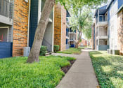 Thumbnail 15 of 32 - a sidewalk between two apartment buildings with grass and trees at The Manhattan Apartments, Dallas, TX