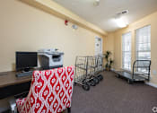 Thumbnail 19 of 25 - A room with a desk, a chair with a red pattern, a printer, a small plant, and two shopping carts.