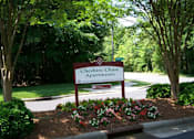 Thumbnail 1 of 6 - A sign for Cheshire Chase Apartments is surrounded by flowers and trees.