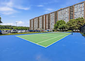 Thumbnail 9 of 20 - A tennis court with a blue surface and white lines is surrounded by apartment buildings.