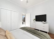 Thumbnail 4 of 32 - A white bedroom with a bed, a television, and a lamp.