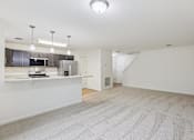 Thumbnail 8 of 48 - A spacious, empty room with a carpeted floor and a kitchen area in the background.