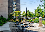 Thumbnail 10 of 12 - A patio with tables and chairs is surrounded by greenery and a building.