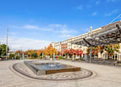Thumbnail 3 of 16 - A fountain in the middle of a plaza with buildings and trees in the background.