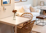 Thumbnail 4 of 6 - A white dining table with a wicker chair and a brown leather ottoman.