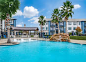 Thumbnail 16 of 17 - A swimming pool surrounded by palm trees and a building in the background.