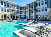 Thumbnail 7 of 27 - A swimming pool with sun loungers in front of apartment buildings at The Grand at Manor Apartments, Manor, TX, 78653