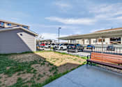 Thumbnail 69 of 70 - A bench is situated on a concrete area with a grassy area and a building in the background.