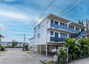 Thumbnail 1 of 23 - A white residential building with blue balconies is surrounded by greenery and other buildings.