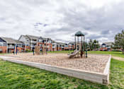 Thumbnail 15 of 20 - A playground with a sandbox and a slide in the foreground and apartment buildings in the background.