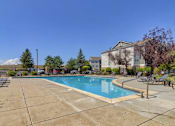 Thumbnail 7 of 20 - A swimming pool surrounded by a concrete patio and a building in the background.