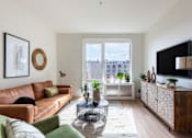 Thumbnail 4 of 67 - A living room with a brown leather couch, a glass coffee table, and a flat-screen TV mounted on the wall.