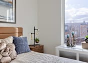 Thumbnail 56 of 67 - A bedroom with a bed, a nightstand with a plant, and a window overlooking a cityscape.