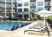 Thumbnail 14 of 20 - A pool with lounge chairs and umbrellas in front of apartment buildings.