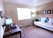 Thumbnail 6 of 18 - A living room with a couch, a desk with a lamp, and a chair.