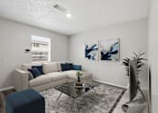 Thumbnail 1 of 22 - A living room with a white couch, a blue ottoman, a glass coffee table, and two abstract paintings on the wall.