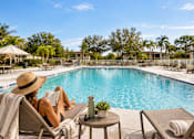Thumbnail 1 of 52 - A woman in a straw hat is reading a book by the pool.