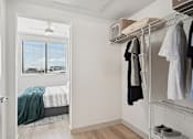 Thumbnail 12 of 26 - A white bedroom with a bed, a window with blinds, a clothing rack with clothes, and a pair of shoes on the floor.