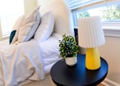 Thumbnail 12 of 27 - A bed with a white comforter and a small potted plant on a table next to a yellow lamp.