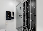 Thumbnail 23 of 35 - A bathroom with a toilet, a glass shower stall, and a black and white tiled wall.