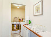 Thumbnail 14 of 23 - A kitchen with a white oven and microwave, a white countertop with a plant, and a framed picture on the wall.