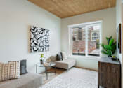 Thumbnail 3 of 15 - A living room with a grey couch, a coffee table, a plant, and a painting on the wall.