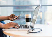 Thumbnail 17 of 19 - A person is using a laptop and holding a smartphone while sitting at a table with a cup of coffee.