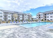 Thumbnail 3 of 41 - A large swimming pool surrounded by a tiled floor and lounge chairs in front of apartment buildings.