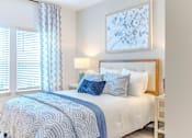 Thumbnail 27 of 41 - A bed with a white comforter and blue pillows is in a room with a white wall and a picture frame.