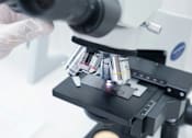 Thumbnail 45 of 45 - A microscope with a slide of samples being held by a gloved hand.