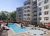 Thumbnail 9 of 38 - A large swimming pool surrounded by lounge chairs and umbrellas in front of a multi-story apartment building.
