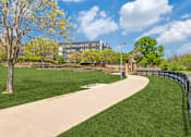 Thumbnail 9 of 40 - A park with a paved walkway, a bench, and a fence.