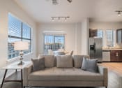 Thumbnail 9 of 20 - A modern living room with a grey couch and a table with a lamp on it.