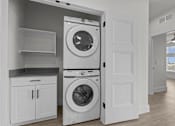 Thumbnail 16 of 24 - A white washing machine is in a laundry room with a cabinet and shelves.