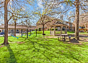 Thumbnail 28 of 30 - A playground with a swing set and a picnic table is surrounded by trees.