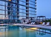Thumbnail 18 of 19 - A modern building with a glass facade reflects the sky, and in front of it is a pool with lounge chairs.