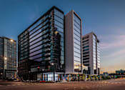 Thumbnail 25 of 26 - A modern glass building stands tall among other buildings at dusk.