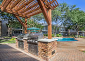 Thumbnail 9 of 24 - A wooden pergola over an outdoor kitchen with a grill and a pool in the background.